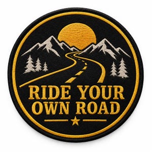 May include: A circular embroidered patch with a black background and a yellow border. The design features a winding road leading towards white mountains and a yellow sun. Pine trees flank the road. The text RIDE YOUR OWN ROAD sits above a star.