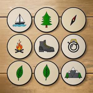 May include: Nine circular coasters featuring outdoor-themed designs. Illustrations include a sailboat, fir tree, compass, campfire, hiking boot, camera, two green leaves, and mountains with trees. Each coaster has a black border.
