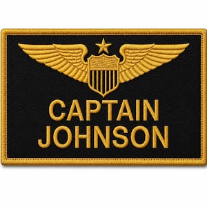 May include: A rectangular black embroidered patch with a gold border. The design features gold embroidered wings flanking a central shield topped with a star. The text CAPTAIN JOHNSON is embroidered in gold capital letters below the emblem.