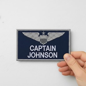Pode incluir: Um patch azul marinho com uma borda prateada, apresentando um emblema prateado de asas e um escudo com uma estrela. O texto "CAPTAIN JOHNSON" está em branco. O patch é segurado por uma mão.