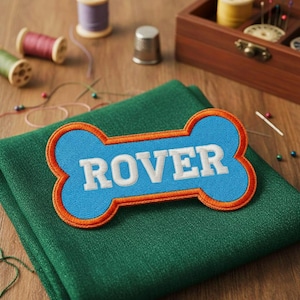 Può includere: Una toppa blu a forma di osso con il nome "ROVER" ricamato in bianco. La toppa ha un bordo arancione ed è posta su un tessuto verde. Nel retro ci sono forniture per cucire, tra cui rocchetti di filo e un ditale.
