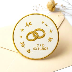 Puede incluir: Invitación de boda bordada con anillos dorados, estrellas y hojas sobre un fondo circular blanco. El texto "C + D 03.11.2027" está bordado en dorado. La invitación tiene un borde dorado y está sobre un sobre crema.