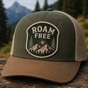 May include: An olive green trucker hat with a brown brim and tan mesh back. A central patch features the text ROAM FREE above an embroidered graphic of a sun, two pine trees, and mountain peaks in shades of green, brown, and yellow thread.