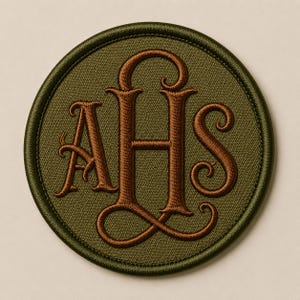 Op de afbeelding: Een ronde geborduurde patch met een olijfgroene achtergrond en een donkerbruine gestikte rand. In het midden staan de letters AHS in een decoratief, in elkaar grijpend schreeflettertype, ook geborduurd met donkerbruine draad.