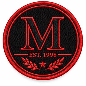 May include: A circular embroidered patch featuring a large red letter M on a black background. Below the letter, the text EST. 1998 is centered above a small red star flanked by two symmetrical red laurel leaf branches, all enclosed by a red border.