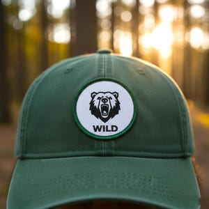 Könnte beinhalten: Eine dunkelgrüne Baseballkappe mit einem runden Aufnäher. Der Aufnäher zeigt eine schwarze Bärenillustration und das Wort "WILD" in Weiß. Die Kappe hat einen gebogenen Schirm und besteht aus einem robusten Material. Der Hintergrund ist verschwommen.