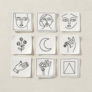 May include: Nine white square coasters with black embroidered designs. The designs include faces, a crescent moon, flowers, a hand holding a flower, and a hand with an open palm. The coasters are arranged on a neutral-colored surface.