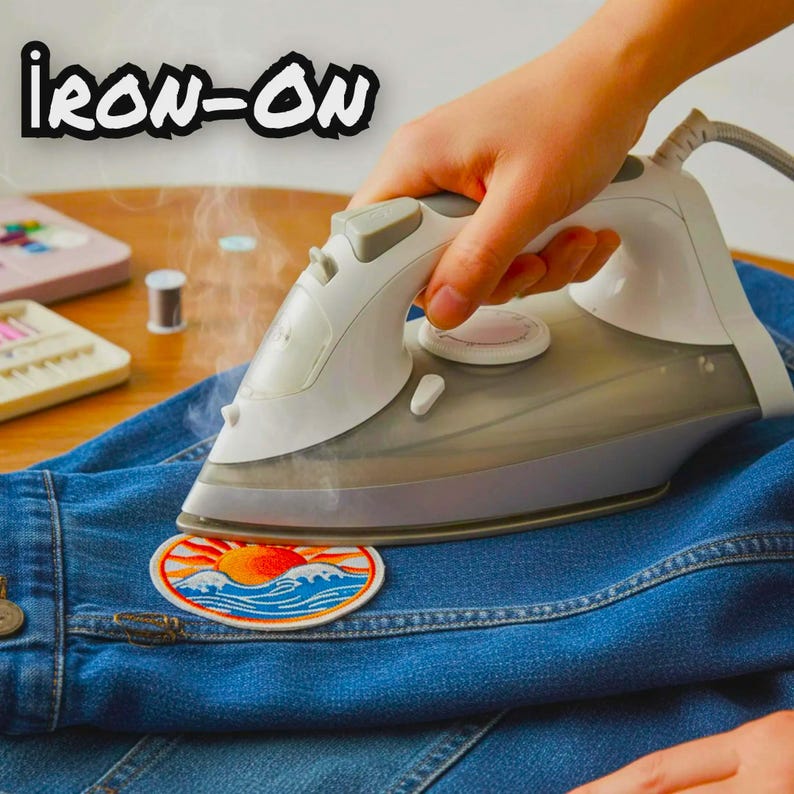 Op de afbeelding: Een wit en grijs stoomstrijkijzer drukt op een ronde geborduurde patch met een gele zon die opkomt boven blauwe oceaangolven, aangebracht op een blauwe spijkerstof. De tekst IRON-ON staat in dikke zwarte letters linksboven.
