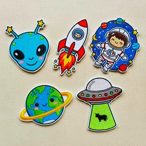 Op de afbeelding: Een verzameling kleurrijke geborduurde patches met ruimte-thema ontwerpen. Inclusief een blauwe alien, een rode en witte raket, een astronaut in de ruimte, een lachende aarde met een gele ring en een UFO met een koe.