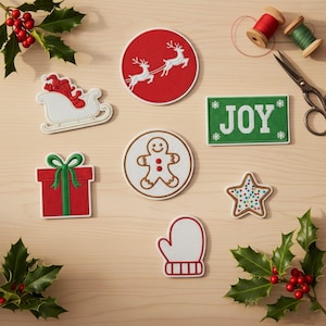 May include: A collection of embroidered Christmas patches, including a sleigh, reindeer, a gift, a gingerbread man, a mitten, a star, and a green sign with the word "JOY". Holly and berries, thread spools, and scissors are also present.