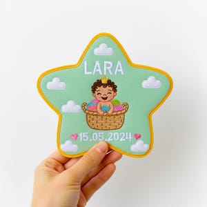 May include: A star-shaped patch with a light green background and a yellow border. The patch features the name "LARA" in white, a baby in a basket of yarn, white clouds, and the date "15.05.2024".