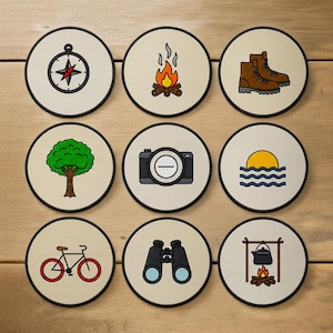 May include: Nine round coasters with camping and outdoor adventure icons. The icons include a compass, campfire, hiking boots, tree, camera, sunset, bicycle, binoculars, and a pot over a fire. Each coaster has a beige background and a black border.