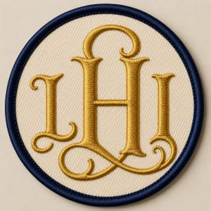 May include: A circular embroidered patch featuring a cream-coloured fabric base with a dark navy blue stitched border. The centre displays a large, ornate monogram in metallic gold thread, consisting of the intertwined letters L, H, and I in a serif font.