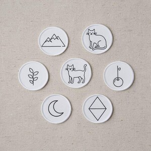 May include: Seven circular white fabric patches featuring black embroidered line art designs. The icons include a mountain range, a sitting cat, a leafy branch, a standing cat, a cherry, a crescent moon, and a diamond shape with a horizontal line.
