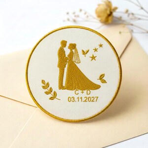 May include: Embroidered wedding patch featuring a bride and groom silhouette in gold thread on a white background. The patch includes the initials "C + D" and the date "03.11.2027". The design is framed by a gold border and includes decorative stars and leaves.