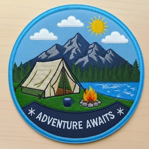 May include: Embroidered patch with a camping scene. A beige tent sits on green grass near a blue lake and forest, with mountains in the background. A campfire and the text "ADVENTURE AWAITS" are also featured. The patch has a blue border.