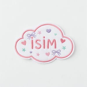May include: Embroidered cloud-shaped patch with the word "ISIM" in pink. The patch features a pink border, hearts, bows, and stars in shades of pink, purple, and green, on a white background.