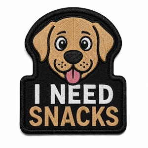Könnte beinhalten: Ein schwarzer bestickter Aufnäher mit einem beigen Hundegesicht und herausgestreckter rosa Zunge. Unter dem Hund ist der Text I NEED SNACKS in weißem und beigem Garn gestickt. Der Aufnäher hat einen dicken schwarzen Rand und eine abgerundete Form.
