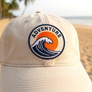May include: A beige baseball cap with a circular patch showing a navy blue wave, an orange sun, and the word "Adventure" in white. The cap has a curved peak and a classic six-panel design.