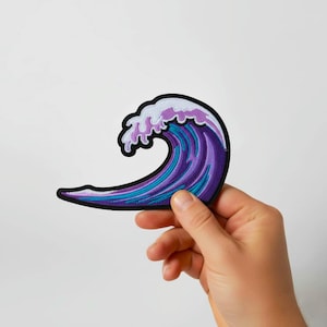 May include: An embroidered patch showing a stylized wave in shades of purple, blue, and white, with a black border. The patch is held against a white background. The design is reminiscent of a Japanese woodblock print.
