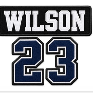 Pode incluir: Um emblema retangular bordado em preto com o texto branco WILSON em fonte de bloco em negrito, posicionado acima de dois grandes números 23 bordados em azul marinho com contornos brancos e pretos, concebidos como emblemas termocolantes.
