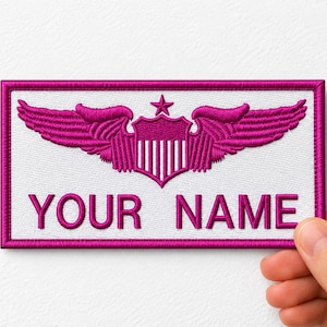 May include: A rectangular embroidered patch with a white background and a magenta border. The design features a magenta winged shield emblem with a star above it, centred above the magenta embroidered text that reads YOUR NAME.
