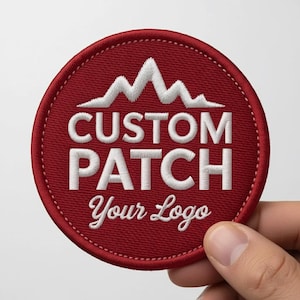 May include: A circular, red embroidered patch featuring white text and a mountain design. The text reads "CUSTOM PATCH" above "Your Logo". The patch is held by a hand against a white backdrop.