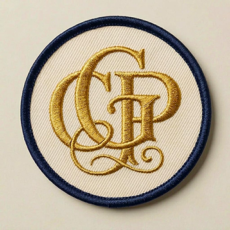 K&ouml;nnte beinhalten: Ein kreisf&ouml;rmiger, bestickter Aufn&auml;her mit einer cremefarbenen Stoffbasis und einem dunkel marineblauen gestickten Rand. In der Mitte befindet sich ein verziertes Monogramm aus metallischem Goldgarn mit den verschlungenen Buchstaben C und P.