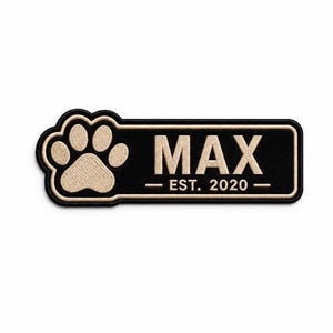 Könnte beinhalten: Ein rechteckiger, schwarz bestickter Aufnäher mit abgerundeten Kanten und einem beigen Pfotenabdruck auf der linken Seite. Der Text MAX ist mittig in Beige gehalten, darunter steht der Text EST. 2020 in einer kleineren beigen Schriftart.