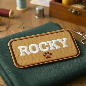 Può includere: Una toppa rettangolare ricamata con sfondo beige e bordo marrone scuro. La toppa presenta il nome ROCKY cucito in lettere maiuscole bianche, posizionato sopra una piccola icona di impronta di zampa ricamata in marrone scuro.