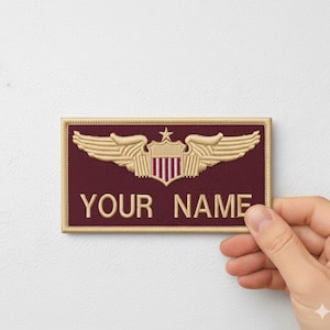 May include: Maroon and gold embroidered patch with the text "YOUR NAME". The patch features a gold emblem with wings, a star, and a shield design. The patch is rectangular and has a gold border.