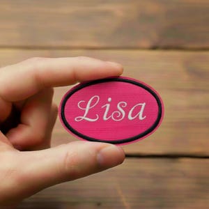 Peut inclure: Un écusson ovale rose avec le nom "Lisa" brodé en blanc. L'écusson a une bordure noire et est tenu dans une main sur un fond en bois. L'écusson mesure environ 7.6 cm de long.