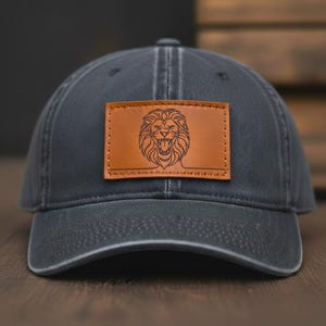 May include: A dark gray baseball cap with a brown leather patch featuring a detailed lion's head design. The cap has a curved brim and a stitched seam running down the center.
