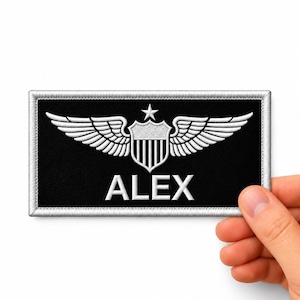 Puede incluir: Un parche rectangular negro bordado con un borde plateado. El diseño presenta un escudo central con rayas verticales, rematado por una estrella de cinco puntas y flanqueado por dos alas extendidas. El nombre ALEX está bordado en letras mayúsculas blancas.