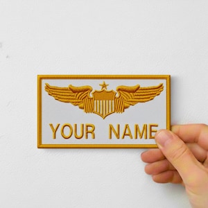 Könnte beinhalten: Gestickter Aufnäher mit goldenen Flügeln, einem Stern und einem Schilddesign, mit dem Text "YOUR NAME" in Gold. Der Aufnäher hat einen weißen Hintergrund und einen goldenen Rand.