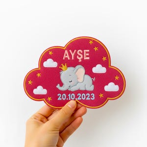 Puede incluir: Parche bordado en forma de nube roja con el nombre "AYŞE" en rosa. Presenta un elefante gris durmiente con una corona dorada, nubes blancas y estrellas amarillas. La fecha "20.10.2023" está bordada en azul.