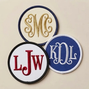 May include: Three circular embroidered patches with monogrammed letters. One patch is cream with gold stitching and a navy border, one is white with red stitching and a black border, and one is blue with white stitching and a white border.