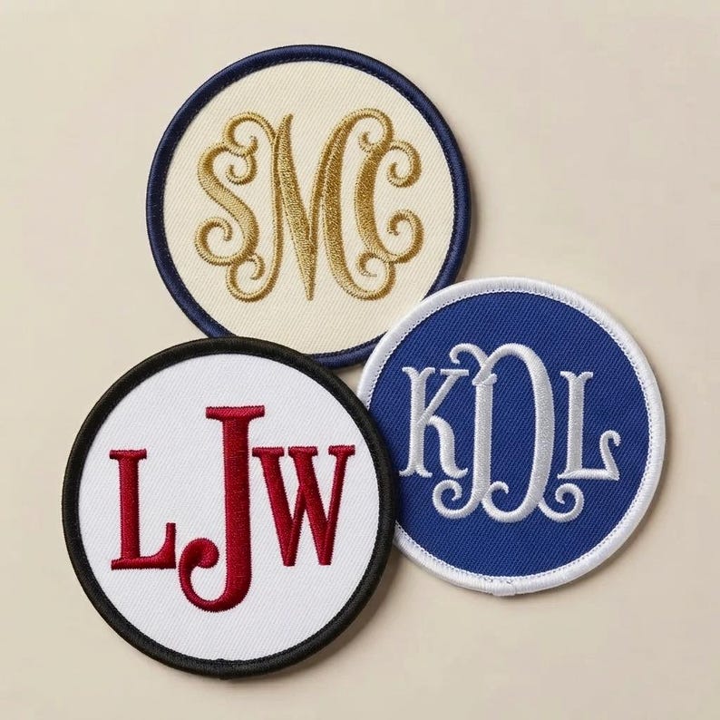 May include: Three circular embroidered patches with monogrammed letters. One patch is cream with gold stitching and a navy border, one is white with red stitching and a black border, and one is blue with white stitching and a white border.