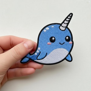 Puede incluir: Parche bordado con un narval de dibujos animados. El parche tiene un cuerpo azul con detalles blancos, una barriga blanca y un cuerno en espiral. El narval tiene una cara sonriente con mejillas rosadas y ojos negros grandes.