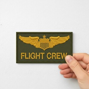 May include: An olive green embroidered patch with gold-colored wings and a shield design. The text "FLIGHT CREW" is embroidered in gold. The patch is rectangular and has a textured appearance.