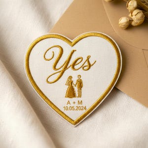 Puede incluir: Parche bordado en forma de corazón con borde dorado y la palabra "Yes" en escritura elegante. Presenta una ilustración de novios y la fecha "10.05.2024". Ideal para recuerdos de boda o anuncios.