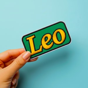 Peut inclure: Un écusson rectangulaire vert avec le nom "Leo" brodé en jaune, bordé de noir. L'écusson est présenté sur un fond bleu clair.