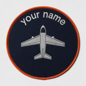 Pode incluir: Um emblema bordado circular com fundo azul-marinho escuro e borda laranja brilhante. O centro apresenta um gráfico de avião cinza claro, e o texto "your name" está bordado em letras cinza claro acima do avião.