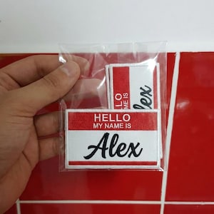 Peut inclure: Un ensemble d'écusson de nom dans une pochette en plastique transparent. Les écussons sont blancs avec une bordure rouge et comportent le nom "Alex" en écriture noire. Un écusson indique "HELLO MY NAME IS" au-dessus du nom.