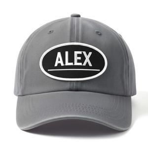 Puede incluir: Gorra de béisbol gris con visera curva. Presenta un parche ovalado negro con el nombre "ALEX" en letras blancas. Un borde blanco rodea el parche, con una línea blanca debajo del nombre.