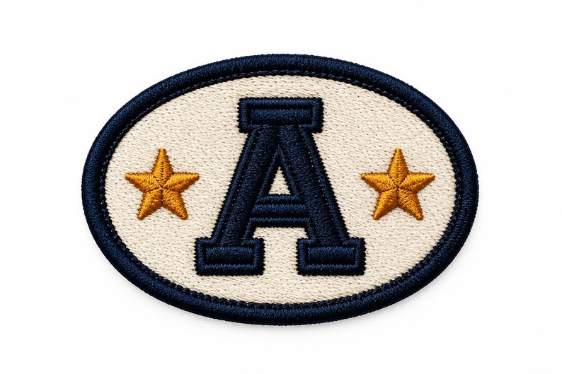 May include: An oval-shaped embroidered patch featuring a cream-coloured background with a dark navy blue border. In the centre, a large dark navy blue letter A is flanked by two five-pointed gold stars, one on each side of the letter.