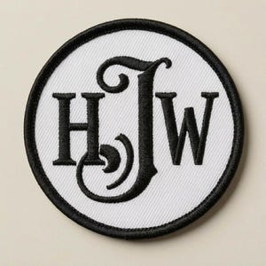 May include: A circular embroidered patch featuring a white background with a thick black stitched border. The centre displays the black embroidered letters H, J, and W, with the letter J designed in a larger, stylised, decorative script font.