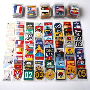 May include: A collection of various embroidered patches in clear plastic packaging. The patches feature flags, stars, text, and graphic designs. Colors include red, white, blue, yellow, and black. Some patches have the word "PATCH" on them.