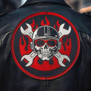 May include: Black leather jacket with a large, round patch featuring a skull wearing a helmet and sunglasses, crossed wrenches, and red flames. The patch has a red border and white and red details.