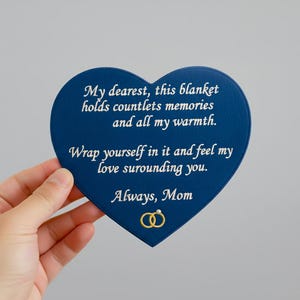 May include: A navy blue heart-shaped blanket with white embroidered text. The text reads, "My dearest, this blanket holds countlets memories and all my warmth. Wrap yourself in it and feel my love surrounding you. Always, Mom."